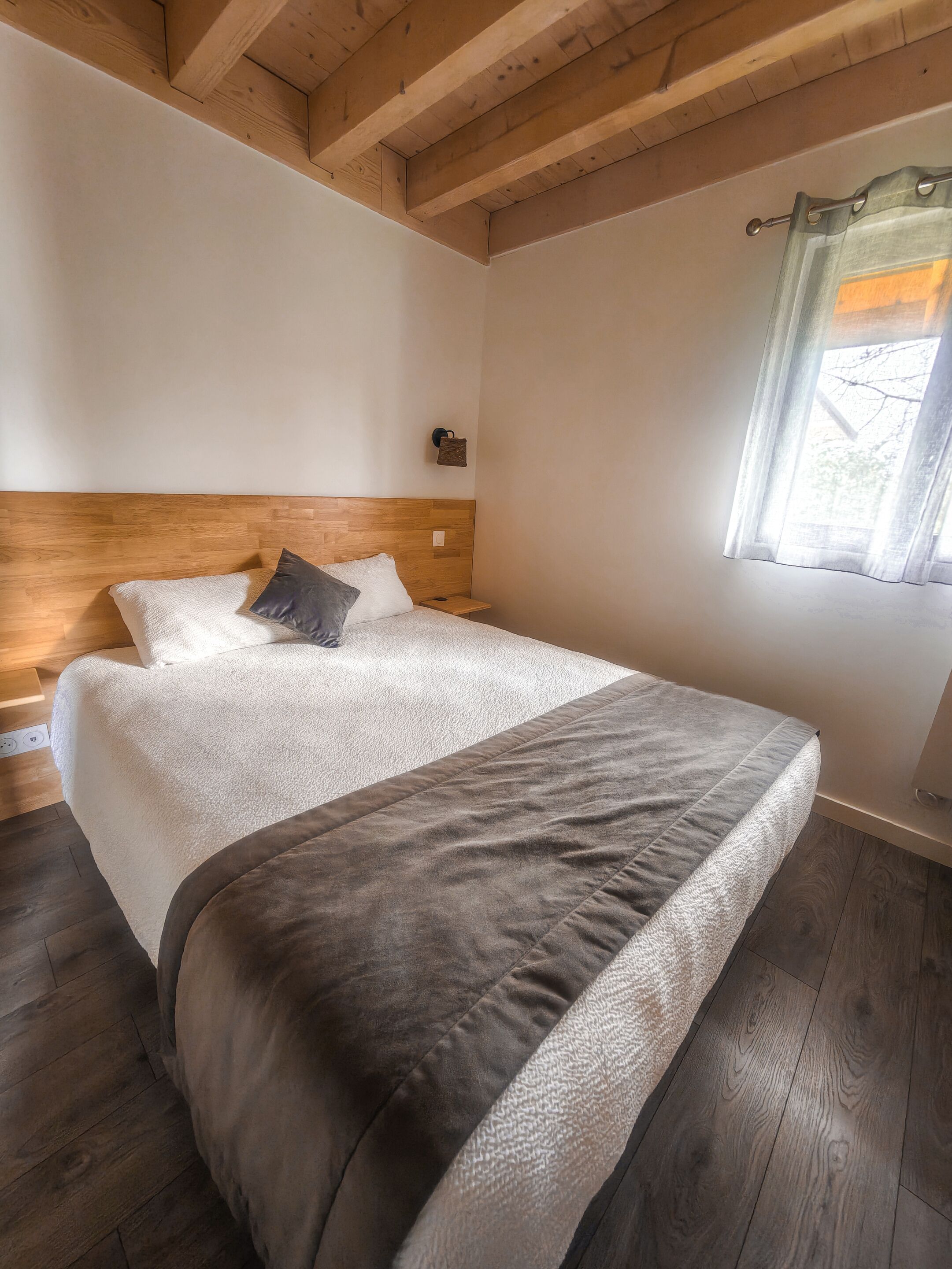 A bedroom with a wooden headboard, a bed with a white and gray blanket, a bedside table, and a window with a curtain.