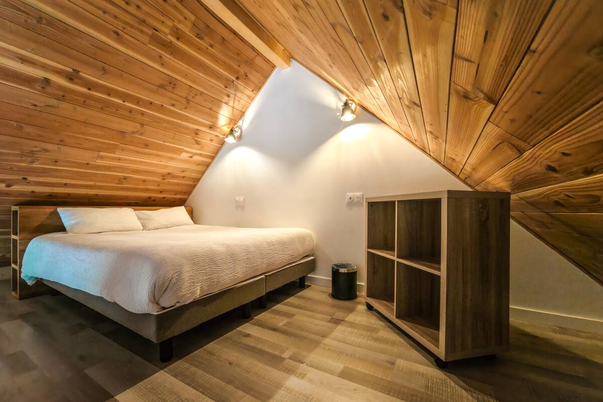 Een knus zolderkamertje met een houten schuine plafond, een bed, een klein houten kastje en een ladder.