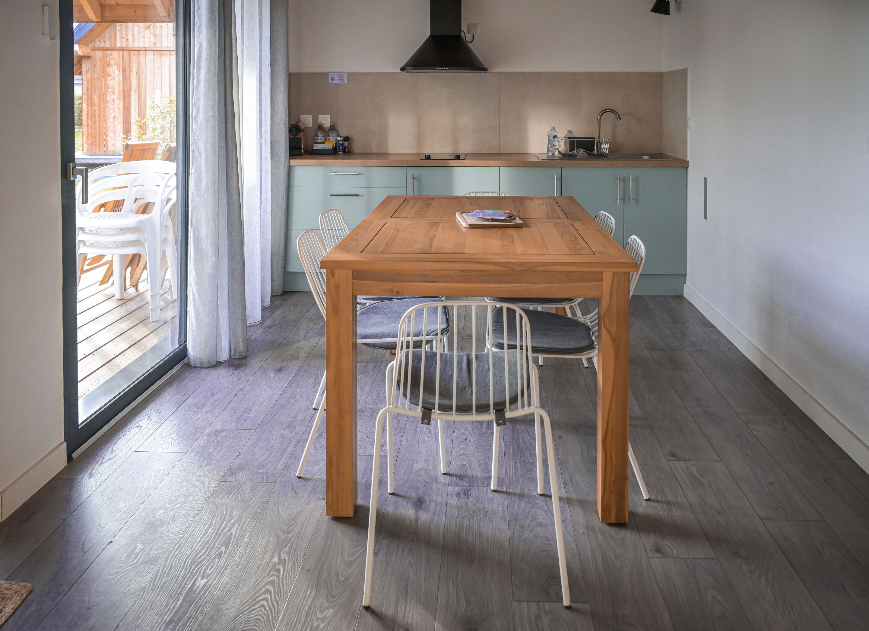 Un coin repas avec une table en bois et des chaises en métal, une cuisine avec des meubles de couleur bleu clair et une vue sur une terrasse en bois, visible à travers une porte vitrée.