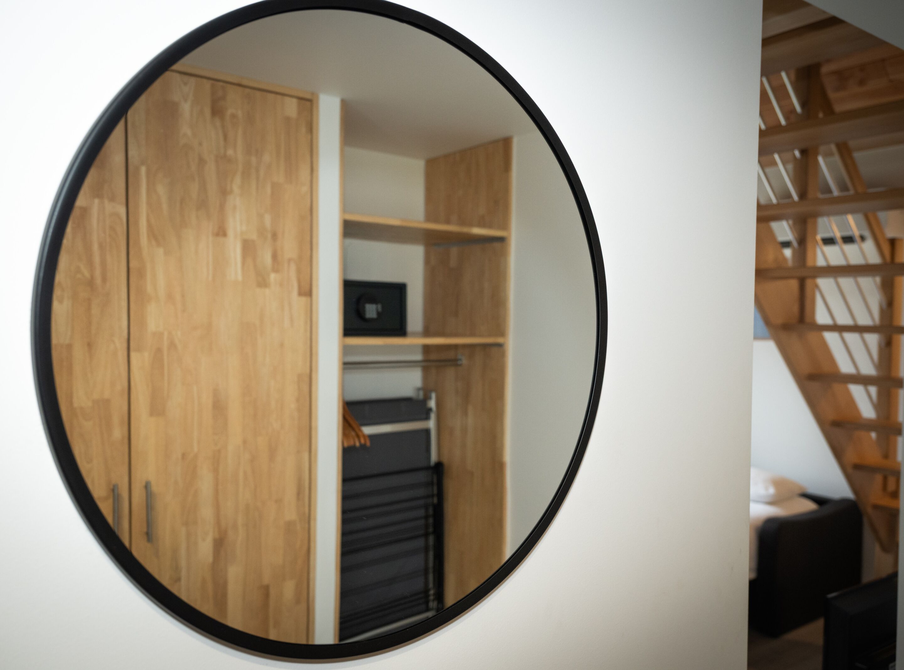 Un miroir rond reflète une pièce avec des meubles en bois, une étagère noire et un escalier avec des rampes en bois.