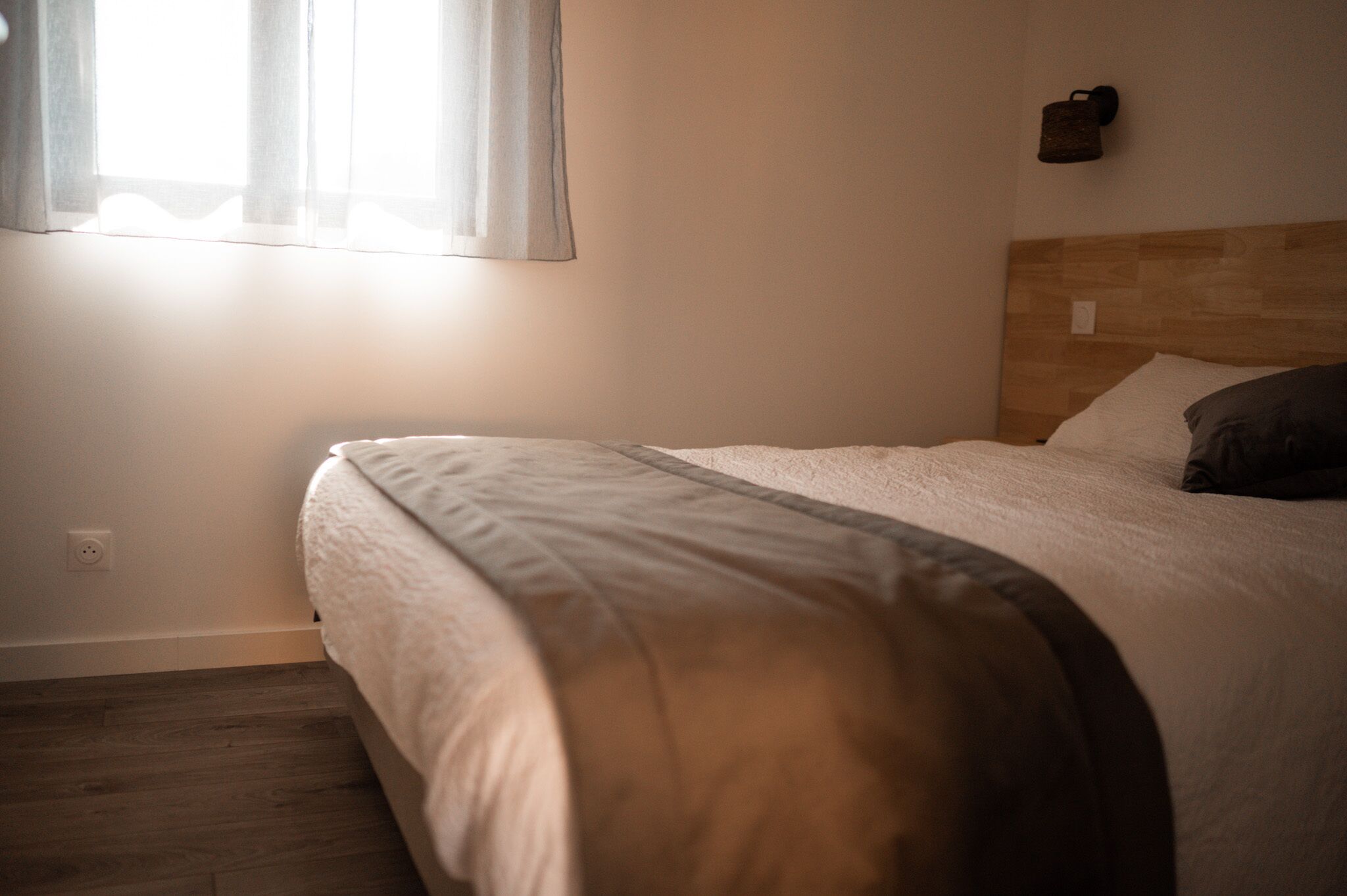 A neatly made bed with a wooden headboard, a bedside lamp, and a window with sheer curtains.