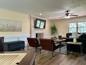 A living room with a brick fireplace, a flat-screen TV mounted on the wall, a ceiling fan, and a large window with a view of trees.