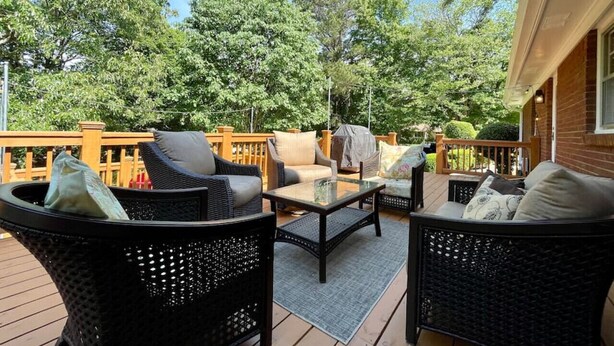 Eine Terrasse im Freien mit Korbmöbeln, einem Tisch mit Glasplatte und Blick auf Bäume sowie einem Geländer aus Holz.