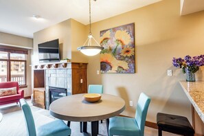 Un coin repas avec une table ronde, des chaises bleues, un téléviseur fixé au mur et un tableau représentant des fleurs.