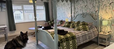 A four-poster bed with a floral wallpaper, a dog sitting on the floor, and a small round table with a lamp.