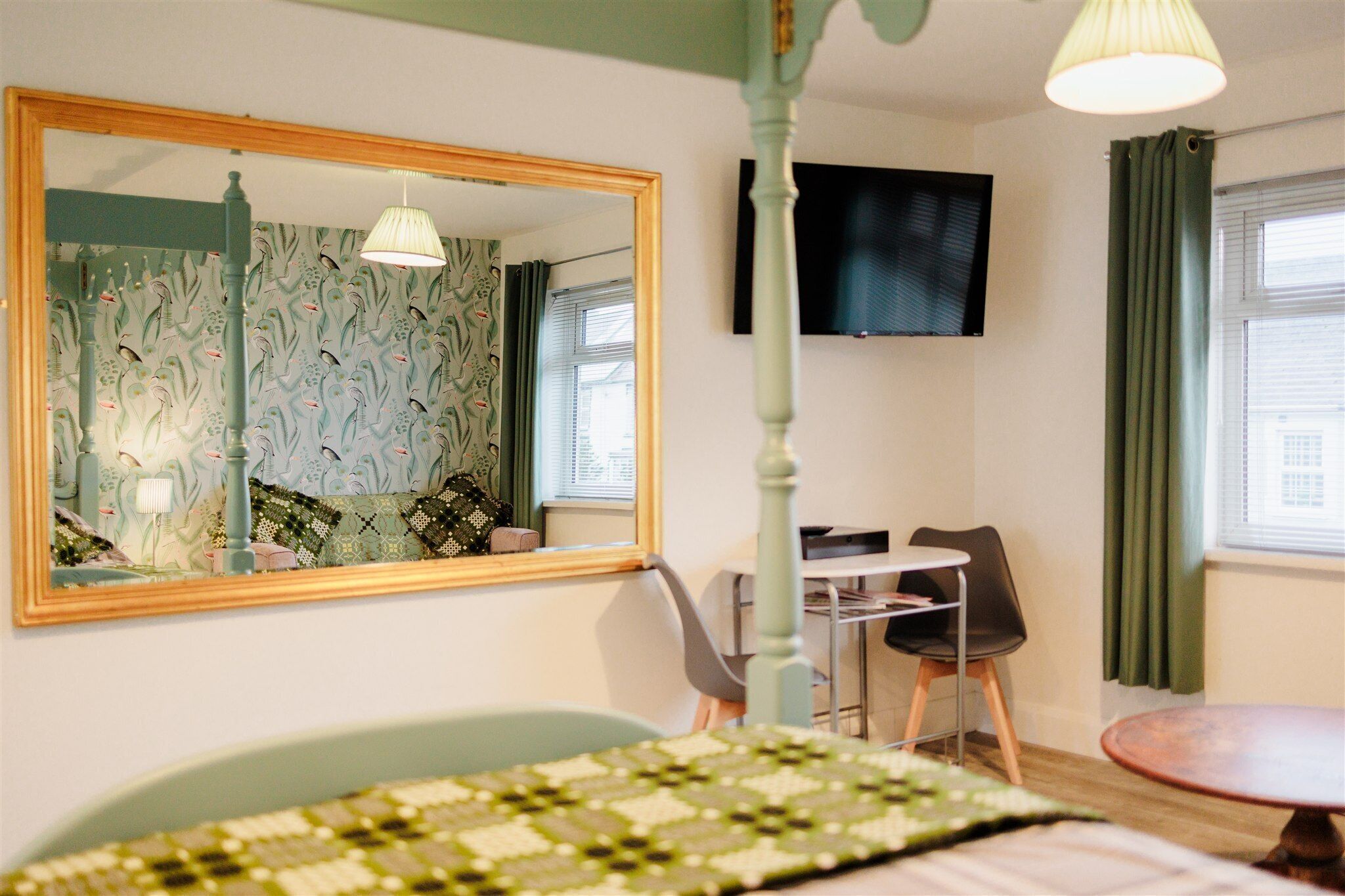 A hotel room with a large mirror, a television mounted on the wall, a dining table with chairs, and a bed with a patterned bedspread.
