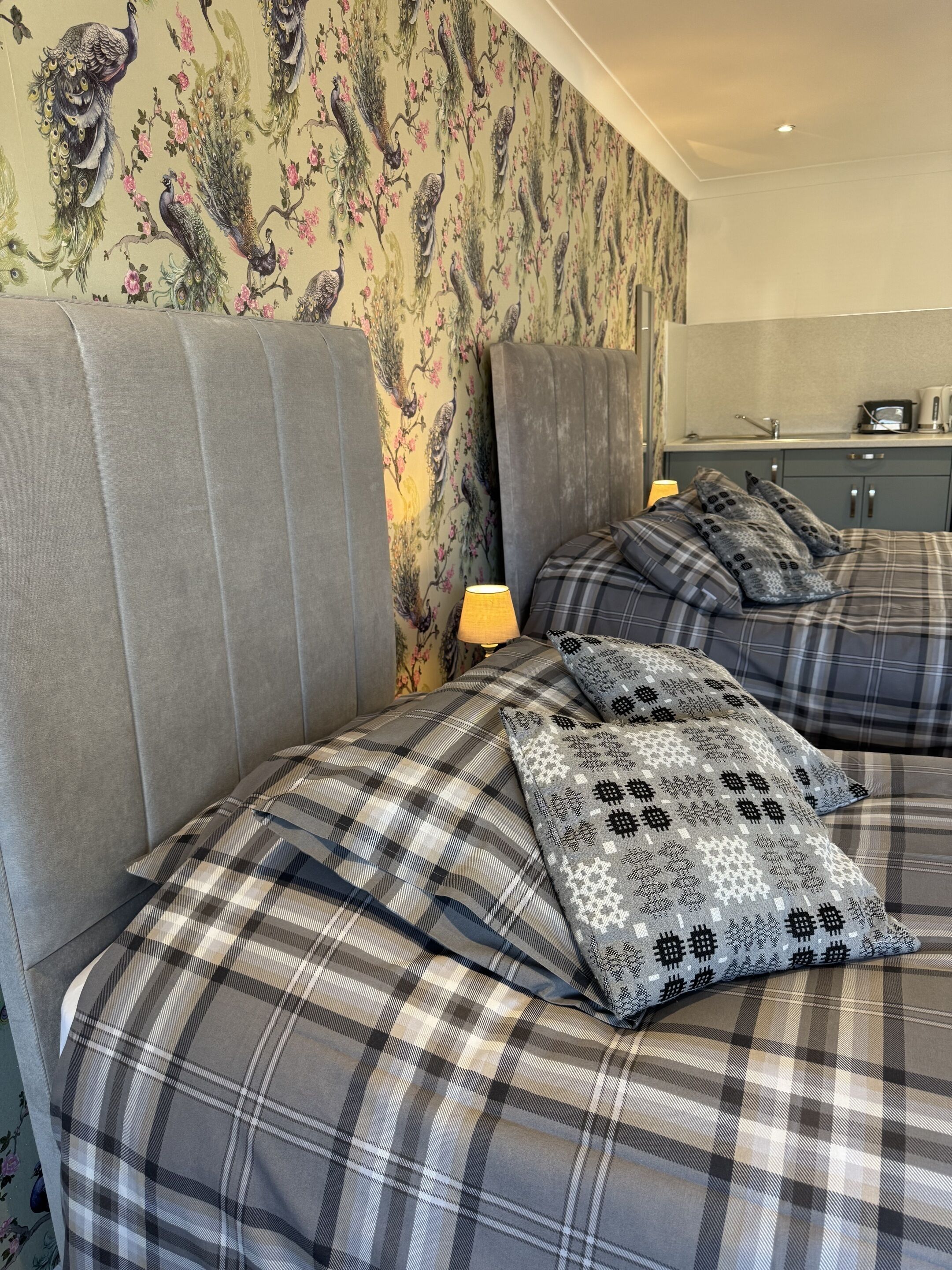 A hotel room with two beds, grey upholstered headboards, and a floral wallpaper.