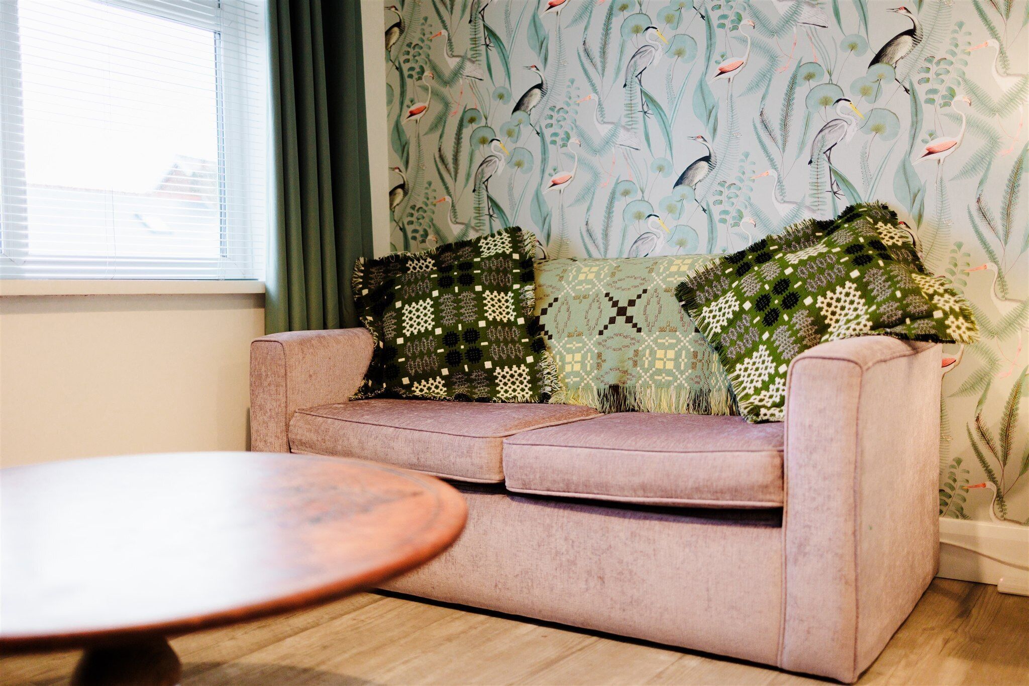 A living room with a sofa, two patterned throw pillows, a round wooden coffee table, and a window with blinds.