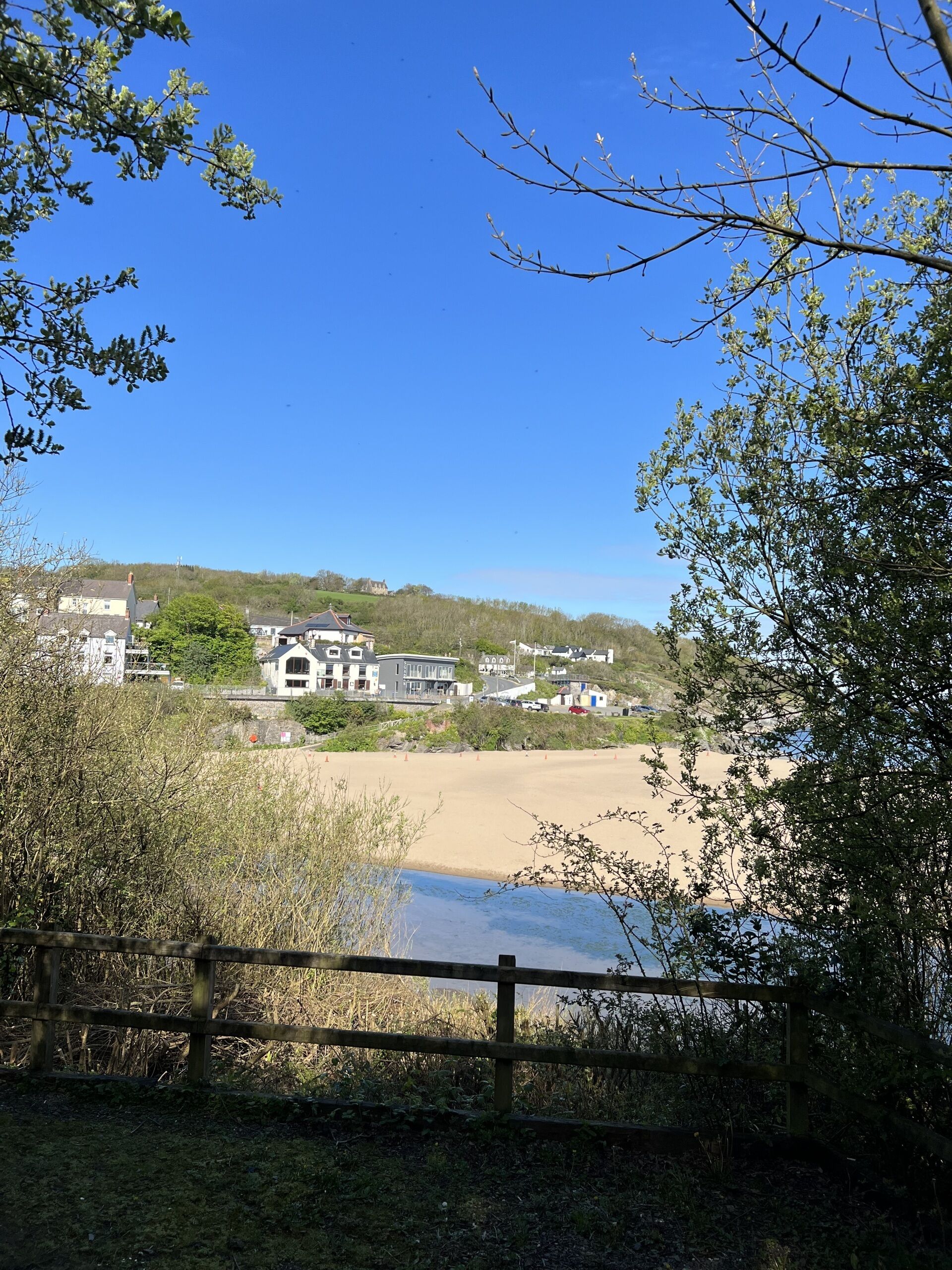 A coastal view with a sandy beach, a wooden fence, and houses in the background.
