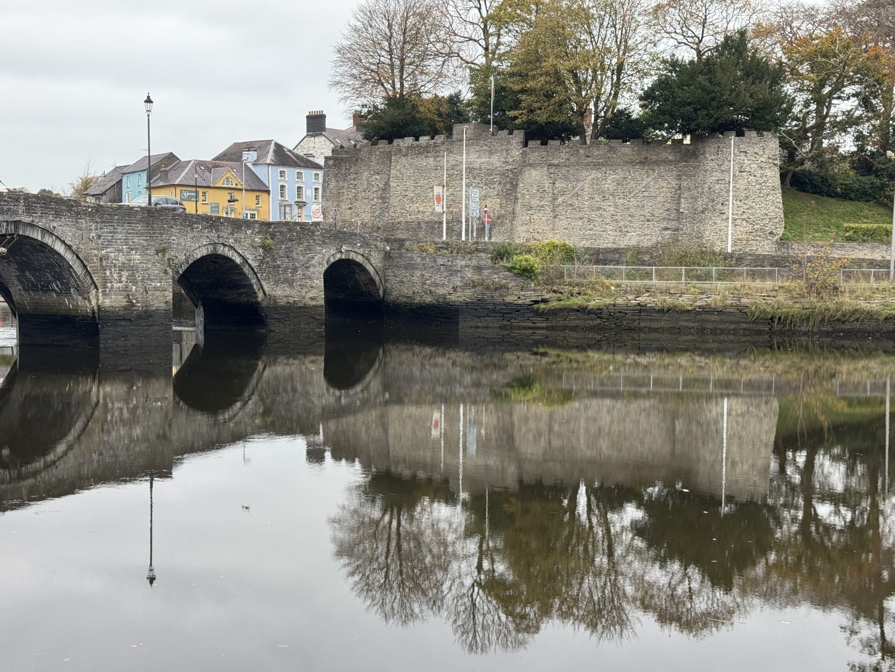 A stone bridge with three arches over a calm river, reflecting the surrounding buildings and trees.