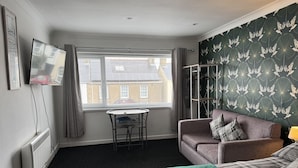 A hotel room with a bed, a sofa, a small table, a TV, and a window with curtains. - Traeth Rooms (Cardigan)