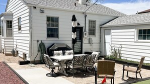 Ein weißes Haus mit einer Terrasse, die einen Tisch, Stühle, einen Grill und einen kleinen Garten mit Kunstgrün und einer Flagge bietet.
