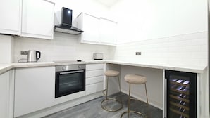 A compact kitchen with white cabinets, a built-in oven, a coffee maker, and two stools. - Dysa Banks Apartments (Blackpool)