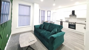 A modern living room with a green sofa, a small coffee table, and a remote control on it. There is a dining area with a table and chairs, and a kitchen with white cabinets and appliances. - Dysa Banks Apartments (Blackpool)