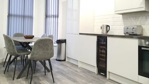 A modern kitchen with white cabinets, a built-in oven, a coffee maker, and a toaster. There is a dining table with chairs and a trash can. - Dysa Banks Apartments (Blackpool)