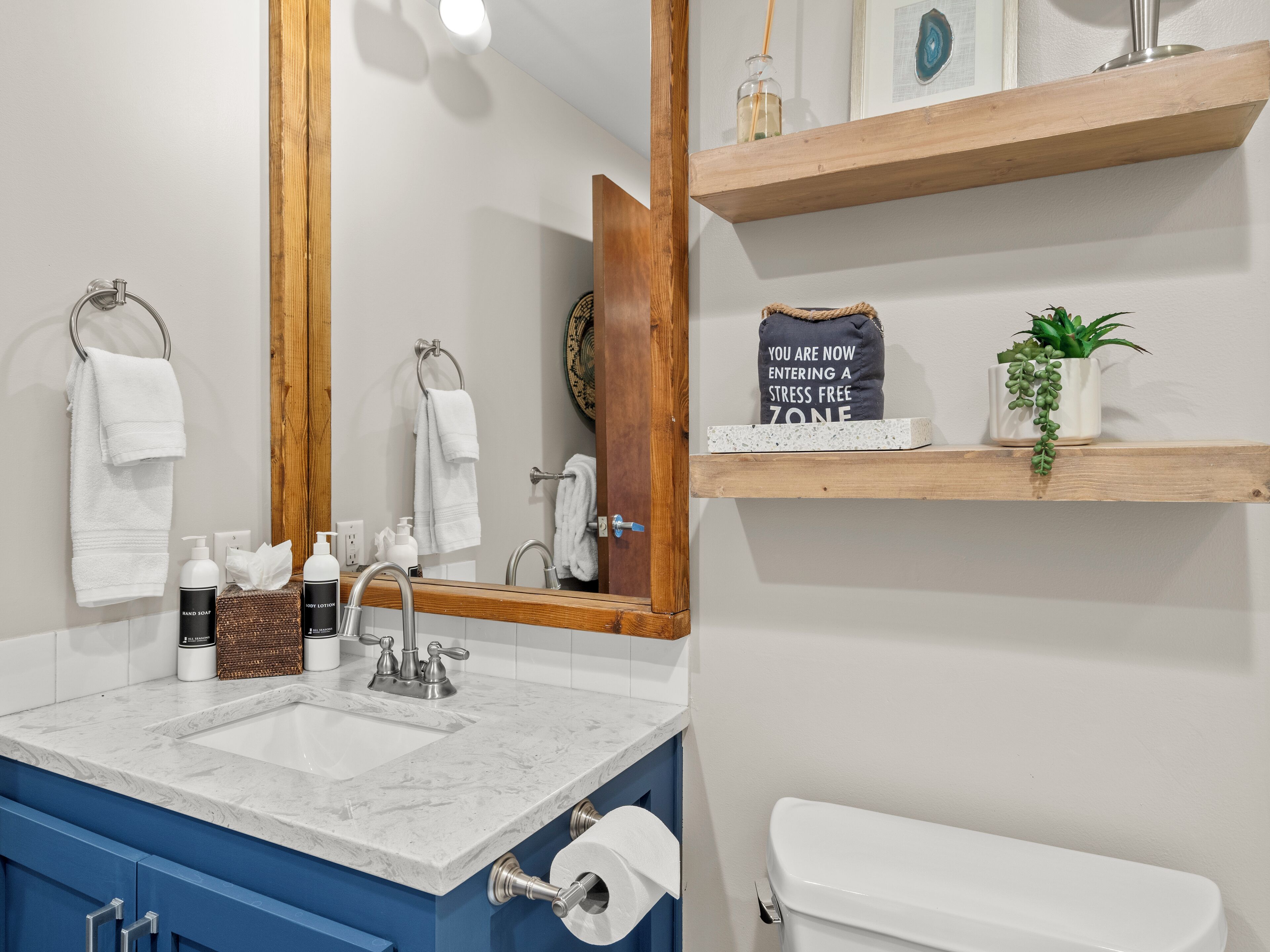 Un baño con lavamanos de mármol, un espejo, un inodoro y repisas de madera con una planta y un cartel.