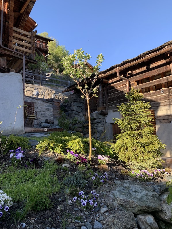A rustic wooden building with a stone wall and a small garden with purple flowers.