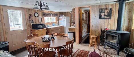 A cozy cabin interior with a dining area, kitchen, and a wood-burning stove.