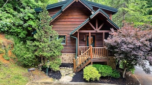 A log cabin with a stone foundation, wooden porch, and a small staircase leading to the entrance.