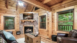 A cozy log cabin interior with a fireplace, leather furniture, and large windows with blinds.