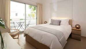 Een moderne hotelkamer met een groot bed, een nachtkastje met een lamp, een stoel en uitzicht op de straat door een groot raam.