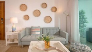 Een moderne woonkamer met een grijze bank, een klein bijzettafeltje met een lamp, een salontafel met een bloevas, en een groot raam met dunne gordijnen.