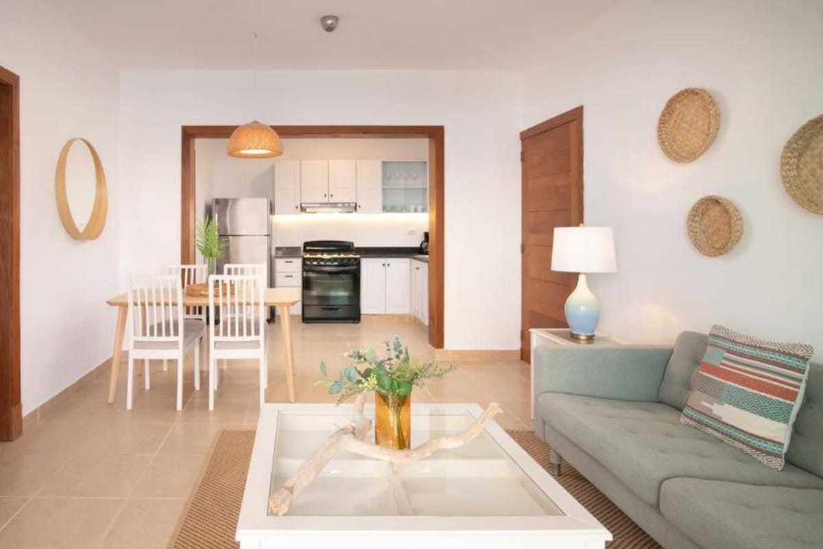 Un salon moderne avec un canapé, une table basse et un téléviseur. On aperçoit une cuisine à l’arrière-plan, avec des placards et une cuisinière.