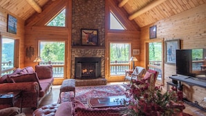 A log cabin interior with a stone fireplace, wooden beams, and large windows offering views of the outdoors. - Crazy Like a Fox Log Cabin | 2 Huge Decks w/ Incredible Views | Pool Table & Foosball (Sparta)