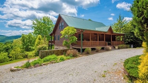 A log cabin with a porch, surrounded by a gravel driveway and lush greenery. - Crazy Like a Fox Log Cabin | 2 Huge Decks w/ Incredible Views | Pool Table & Foosball (Sparta)