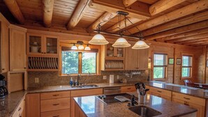 A spacious kitchen with granite countertops, wooden cabinets, and a large island with a sink. There are three pendant lights above the island and a window providing natural light. - Crazy Like a Fox Log Cabin | 2 Huge Decks w/ Incredible Views | Pool Table & Foosball (Sparta)
