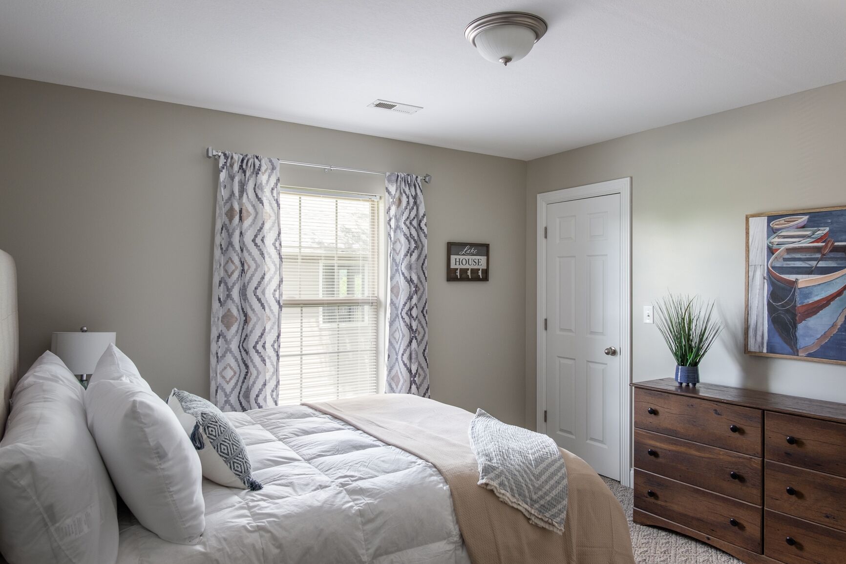 A bedroom with a bed, a dresser, a window with curtains, a door, and a painting on the wall.