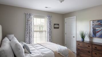A bedroom with a bed, a dresser, a window with curtains, a door, and a painting on the wall.