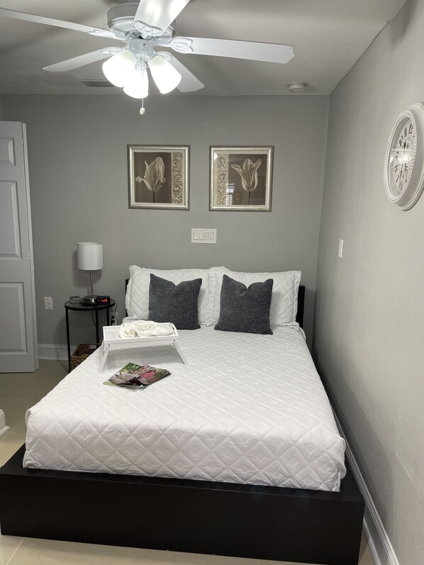 A neatly made bed with a white quilt, two dark pillows, and a tray with a magazine. There is a ceiling fan above, a framed picture on the wall, and a small table with a lamp beside the bed. - La Palma in Port Richey (Port Richey)