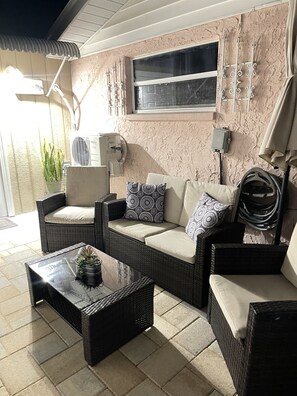 A patio area with wicker furniture, including a sofa and chairs, a glass coffee table with a plant, and a window with a view.
