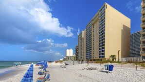 Una playa con una fila de sombrillas azules y sillas reclinables, un edificio alto de color beige y un cielo azul despejado.