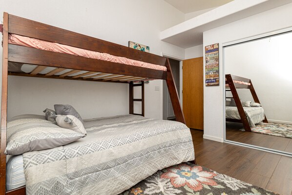 A bunk bed with a wooden headboard, a patterned bedspread, and pillows. There is a small rug on the floor and a poster on the wall.