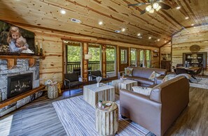 Cozy cabin interior with a stone fireplace, flat-screen TV, and large windows offering a forest view.