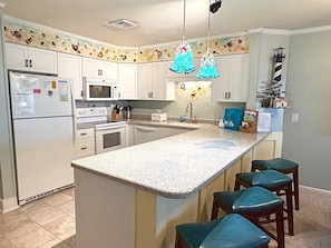 Una cocina con armarios blancos, encimera de granito, microondas, estufa y lavamanos. Hay cuatro taburetes de bar con cojines color turquesa.