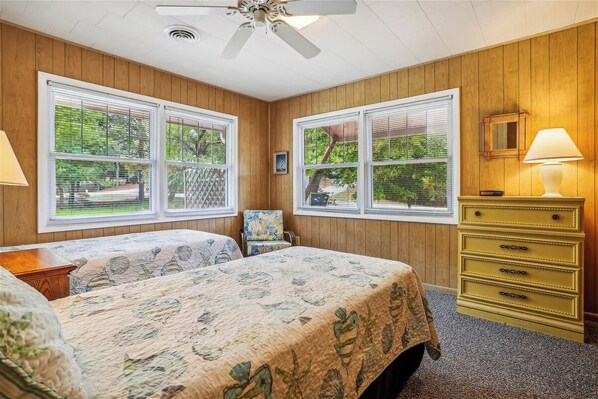 A bedroom with two beds, wooden walls, large windows, a ceiling fan, a dresser, and a lamp. - Seashell - Quaint 3BR Beach Cottage that allows Dogs (Southern Shores)