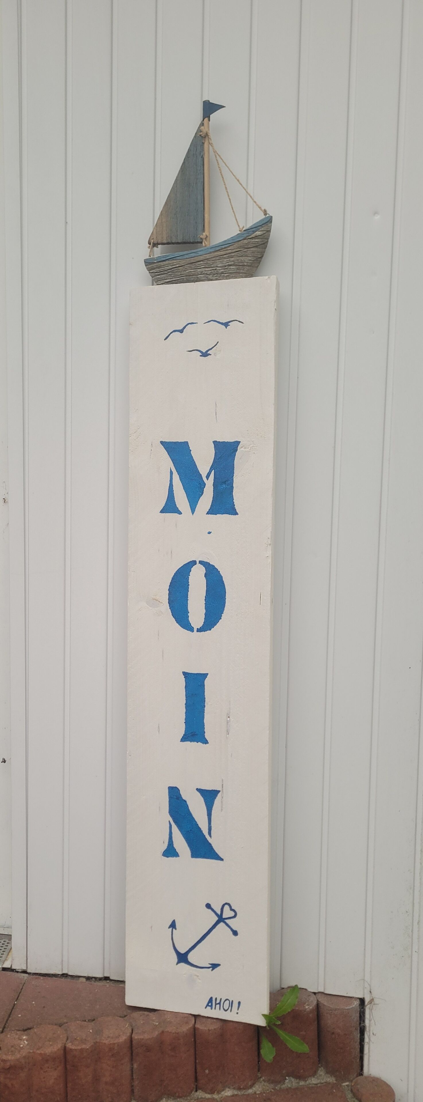 Ein vertikales Schild mit dem Wort 'MOM' in großen, blauen Buchstaben, darüber ein kleines Segelboot und ein Vogel, der nach rechts fliegt.
