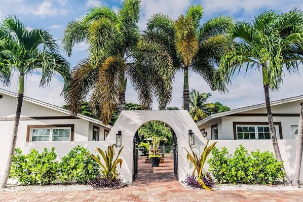 A gated entrance with palm trees and a brick pathway leading to a building with white walls and windows.