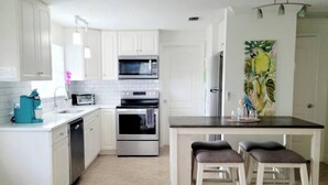 A kitchen with white cabinets, a microwave, oven, and a refrigerator. There are two stools at a table.