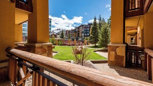 A hotel corridor with wooden railings, a view of a green courtyard, and multi-story buildings in the background. - Sundial C004 | Ski-in/Ski-out | Mountain Views | Private Balcony | Pools & Hot Tubs (Park City)
