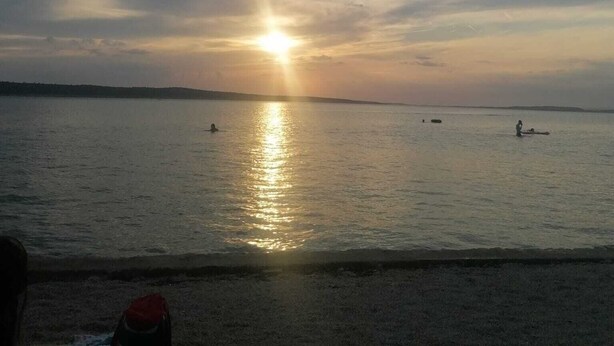 Una spiaggia con il sole che tramonta sull'acqua, mare calmo e alcune persone in lontananza.