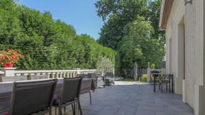 An outdoor patio with chairs and tables, surrounded by lush greenery and a building with a white facade.