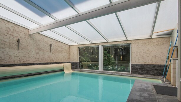 A swimming pool with a glass ceiling, a bench, and a view of greenery outside.