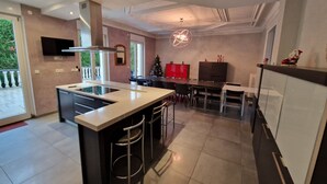 A modern kitchen with a central island, bar stools, a dining area with a table and chairs, a built-in oven, and a TV mounted on the wall.