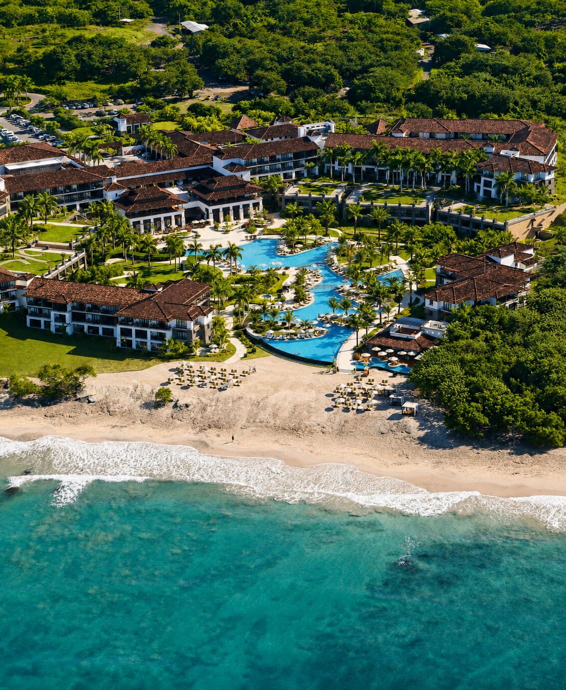 An aerial view of a resort with a central swimming pool, beachfront, and lush greenery.