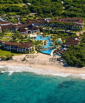 Vista aérea de un resort con piscina central, acceso a la playa y abundante vegetación.