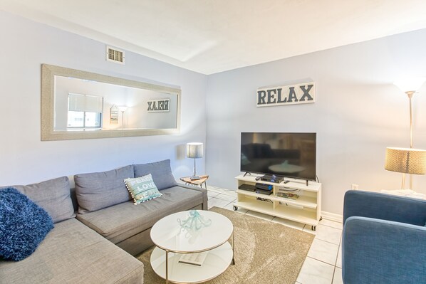 Een moderne woonkamer met een grijze bank, een salontafel, een televisie en een bord met de tekst 'RELAX' aan de muur.